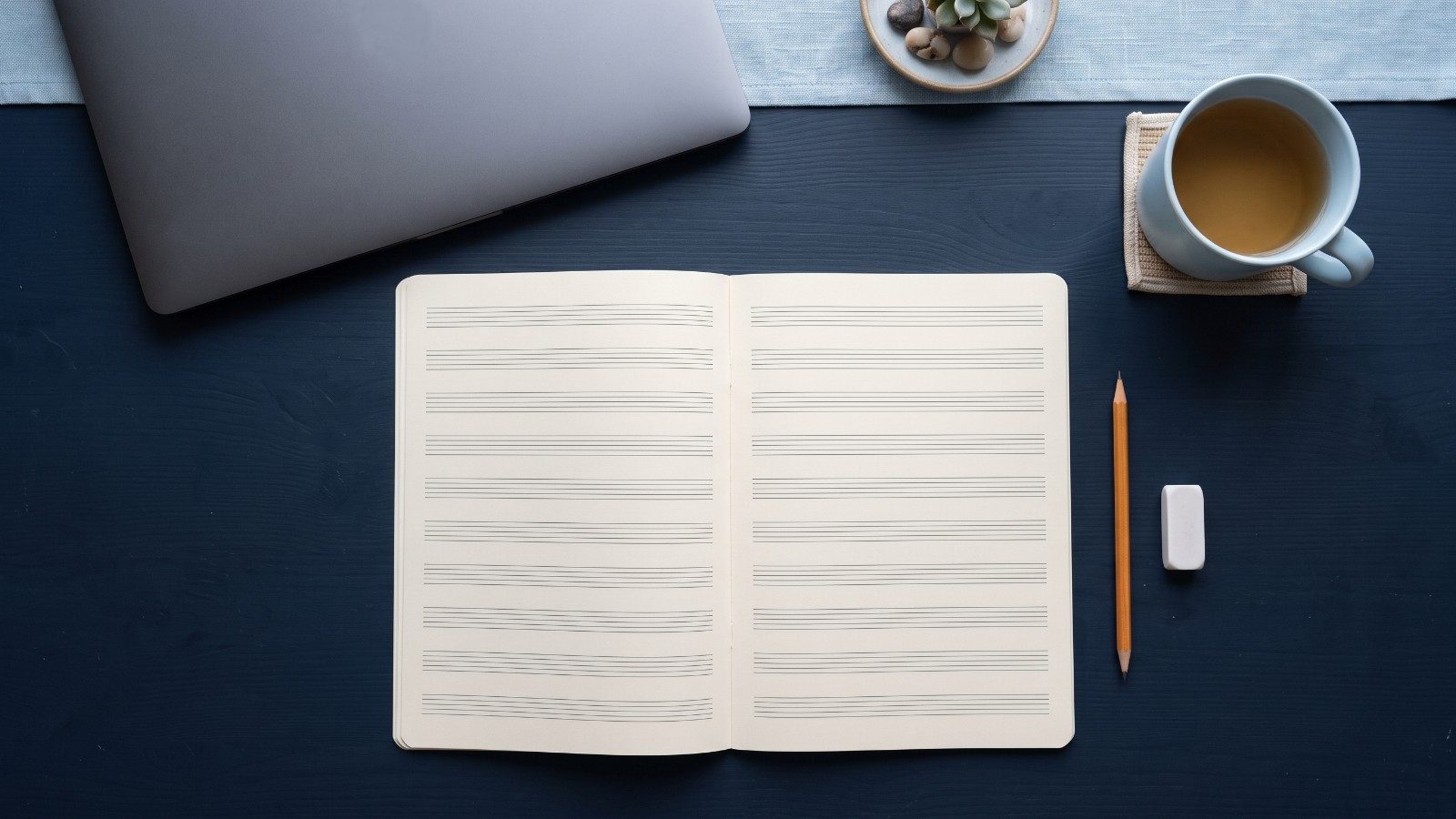 Un cahier de musique ouvert aux portées vides sur un bureau bleu nuit, avec un crayon, une gomme, un ordinateur portable fermé et une tasse de thé