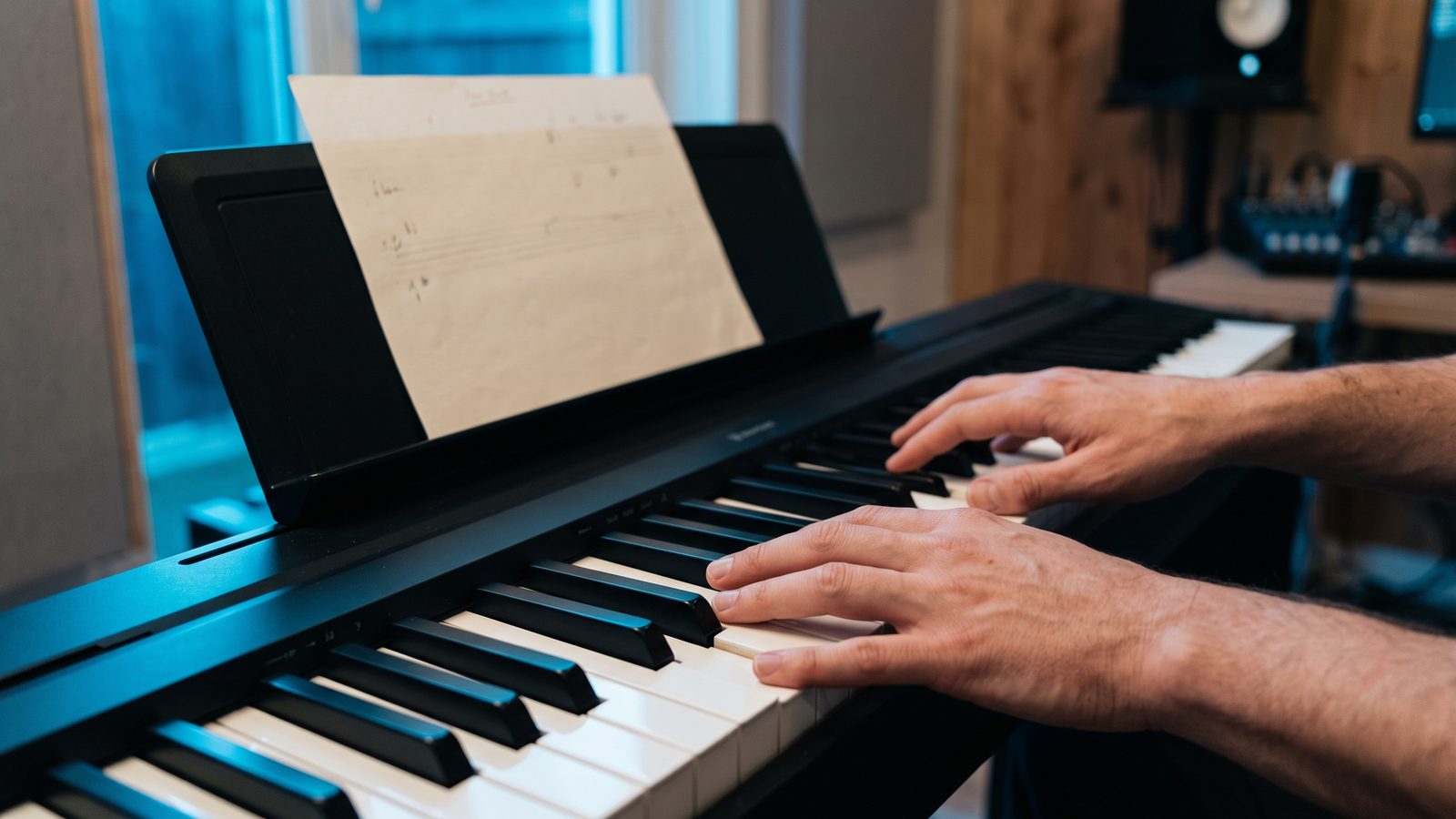 Primer plano de unas manos sobre el teclado de un piano junto a una hoja de acordes escrita a mano en el atril, con luz azul de atardecer